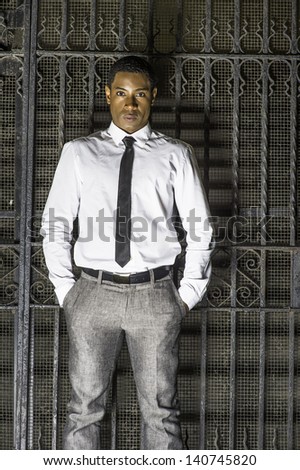 Dressing in a white shirt, a black tie, gray pants, and two hands putting in pockets, a young handsome black businessman is standing in the front of a metal gate and angrily looking at you./Attitude