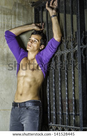 Leaning on the corner between a wall and a metal gate, a sexy young guy is  rolling his purple sweater over his head, showing his well defined body and into deeply thinking./Summer Heat