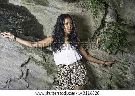 A pretty black girl relies on rocks, with spring water on rocks, stretching arms and thoughtfully looking up./Looking for you