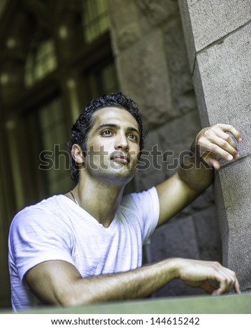 Dressing in white T shirt and two hands touching on window frames, a young handsome guy with bright piercing eyes is thoughtfully looking up/Looking up