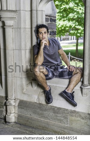 Dressing in a gray polo shirt, pattern shorts and black leather sneakers, one hand touching his chin,  a young strong handsome guy is sitting on a old fashion window frame and looking at you.