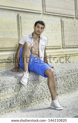 Dressing in a iron short sleeve shirt, blue shorts and sneakers , a young attractive guy is sitting against a pattern wall and relaxing. /Relaxing Outside