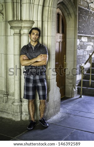 Dressing in a gray short sleeve shirt, pattern shorts and black leather sneakers, a young strong handsome guy is standing by a old fashion wall, crossing arms and thoughtfully looking at you.