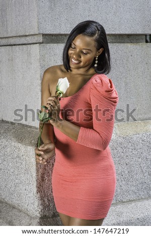 Dressing in a pink one shoulder dress, a black teenage girl is smilingly looking at a white rose. / Black GIrl and White Rose