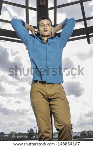 Dressing in a blue shirt, brown pants, two hands putting on the back of the head, a young attractive guy is standing in the front of the broad sky, looking forward / Looking forward