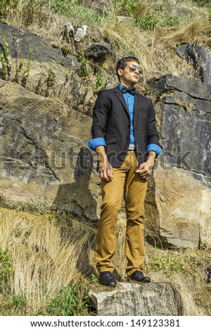 Dressing in a black blazer, a blue shirt, brown pants and a black bow tie,  a young businessman wearing sunglasses is standing on rocks and looking faraway. / Looking Faraway
