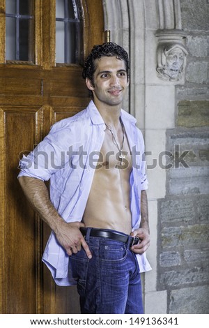 Wearing a cross necklace, with his shirt unbuttoned, a handsome, muscular guy is standing by a old fashion doorway and relaxing. / Relaxing Outside