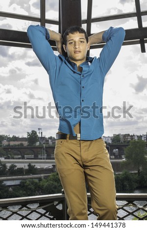 Dressing in a blue shirt, brown pants, two hands putting on the back of the head, a young attractive guy is standing in the front of the broad sky, looking forward. / Looking forward