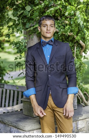 Dressing in a black blazer, a blue shirt, brown pants and a black bow tie,  a young businessman is standing outside, charmingly looking at you. / Portrait of Young Businessman