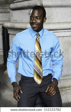 Dressing in a light blue shirt, a colorful pattern tie, a young black businessman is smiling, sitting outside to take a break. / Happy Black Businessman