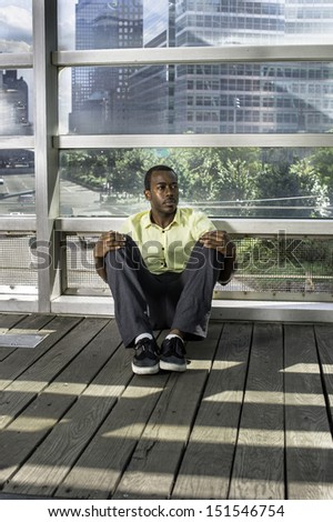 Dressing in a light yellow shirt, gray pants and black shoes,  a young black guy is sitting on the floor by a big glass wall. The background is a busy street scene. / City Life