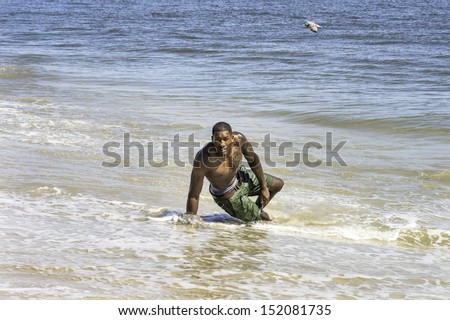 A well built young  black guy, half naked,  is playing with water on the beach. There is a small bird on the background. / Playing on the beach