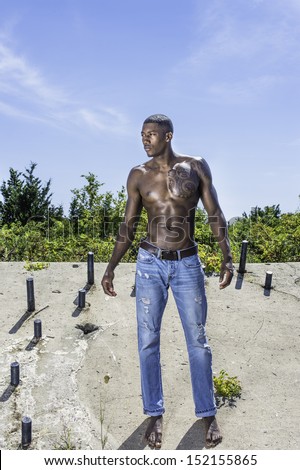 Under the sunshine, a masculine black guy, wearing blue jeans, barefoot and half naked, is standing on a old construction site, confidently looking forward. / Portrait of  Young Black Fitness Guy