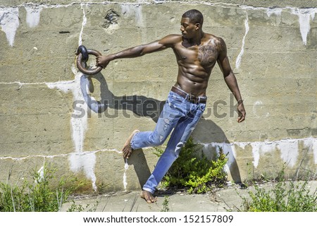 Under the sunshine, a masculine black guy, wearing blue jeans, barefoot and half naked, is standing by a wall and holding a metal ring. / Portrait of  Young Black Fitness Guy