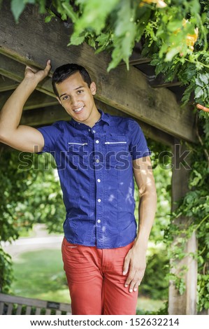 Wearing a blue short sleeve shirt and red pants, a handsome, attractive guy is standing by a wooden structure and green leaves, charmingly looking at you / Portrait of Young Guy