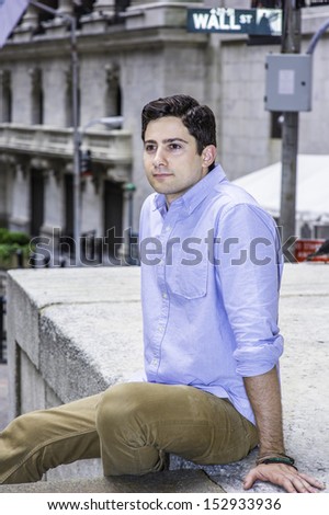 Shrugging his shoulder, a young handsome guy is sitting outside on a stage to relax. There is a Wall Street sign in the background. / Relaxing outside