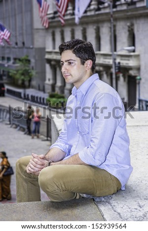 Clamping his hands, a young handsome guy is sitting outside on a stage into deeply thinking. There are American flags hanging on buildings in the background. / Thinking Outside