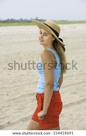 A pretty blonde woman, dressing in light blue tank top and red shorts, wearing a yellow straw hat, is charmingly standing on a sandy beach. / Portrait of Pretty Girl
