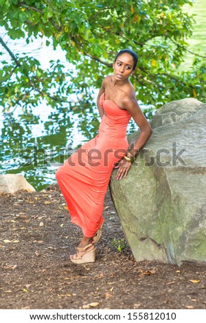 Dressing in a contemporary style red strapless long dress,  a young pretty girl is leaning on rocks by a lake,  relaxing and thinking. / Relaxing Outside