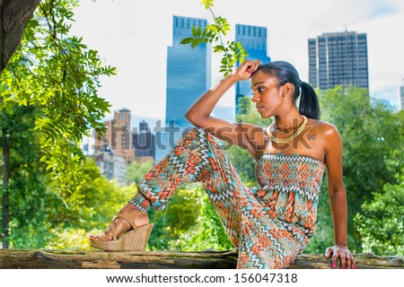 One arm supporting her head, a pretty black girl is sitting outside in a small woods and into deeply thinking. The far background is big city high buildings. / Thinking Outside