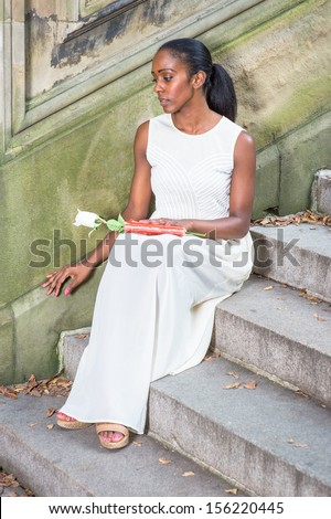 Holding a white rose and a red book on her leg, sitting on steps,  a young pretty black girl is relaxing outside and thinking. / Relaxing Outside