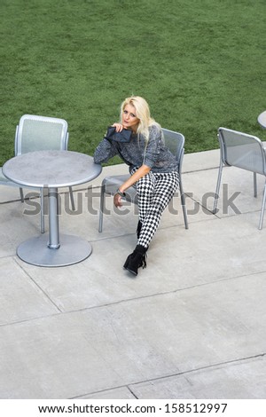 Dressing in a gray fashionable sweater, black and white pattern pants, black boots, holding a black purse, a young blonde girl is sitting on a chair by a lawn, relaxing. / Sitting Down and Relaxing