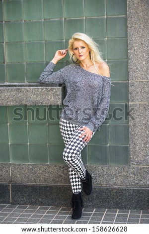 Dressing in a gray fashionable sweater, black and white pattern pants, black boots, a young blonde girl is leaning against a green wall,  relaxing. / City Girl