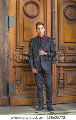 Dressing in a red undershirt, a black blazer, black jeans and a black tie, a young college student is standing in the front of old fashion style door, looking forward. / Portrait of College Student