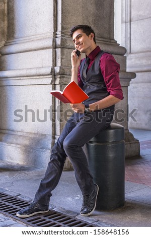Dressing in a red shirt, a black vest, black jeans and a black tie, a young handsome guy is reading a red book while talking on the phone. / Reading Outside