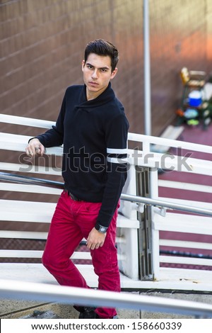 Dressing in a black shawl collar sweater and red jeans, a young handsome guy is standing by a white fence, confidently looking forward. / Portrait of Young Guy