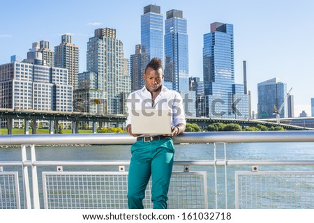 Dressing in a white shirt and green pants,  a young black guy with mohawk hair is standing in the front of high buildings by the water, working on a computer. / Working Outside