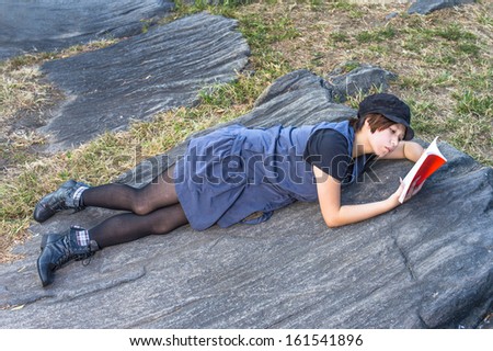 Dressing in a blue sleeveless long blouse, black leggings and boots, wearing a black corduroy cop, a young Chinese girl is lying on rocks, reading a red book. / Reading Outside