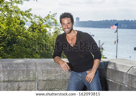 Dressing in a black sweater and blue jeans, a handsome, sexy, middle age guy with mustache and beard is standing against a half wall fence by the river, smilingly looking at you. / Traveler