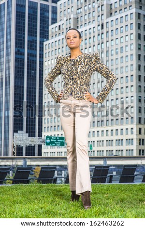 Dressing in black and yellow pattern jacket, light cream colored pants, hands on her hips, a young black businessman is standing in the front of a business district, confidently looking forward.