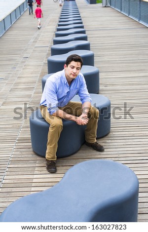 Dressing in a light blue shirt, dark yellow jeans and brown boot shoes, a young handsome guy is sitting on a modern style bench, relaxing and thinking. / Thinking Outside