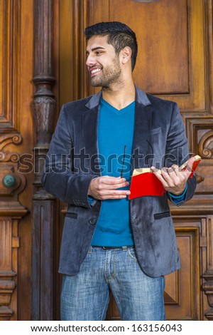 Dressing in a woolen blazer, knit sweater and jeans, holding glasses and a red book,  a young handsome professor with bread and mustache is standing in the doorway, smilingly looking away.