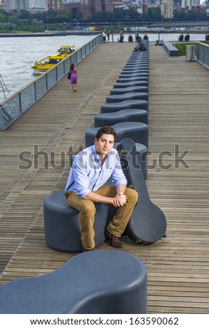 Dressing in a light blue shirt, dark yellow jeans and brown boot shoes, a young handsome musician with an instrument box is sitting on a modern style bench in the evening, charmingly looking at you.