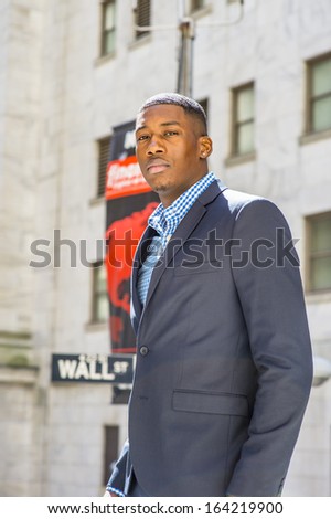A young black businessman is standing outside, confidently looking forward. There is a Wall Street sign in the background. / Portrait of Young Black Businessman