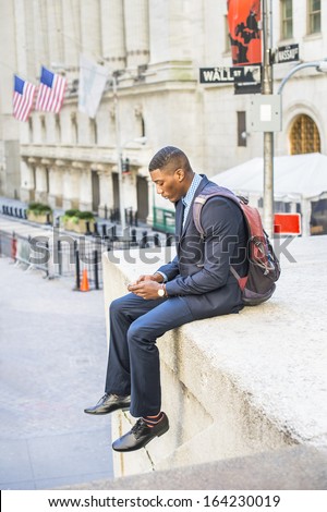 Dressing in a blue suit and leather shoes, caring a backpack, a young black businessman is sitting outside to check messages on his mobile phone. There is a Wall Street sign in the background. / Text