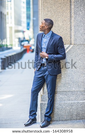 A young black businessman is standing on the street by a column,  looking up. / Looking Up