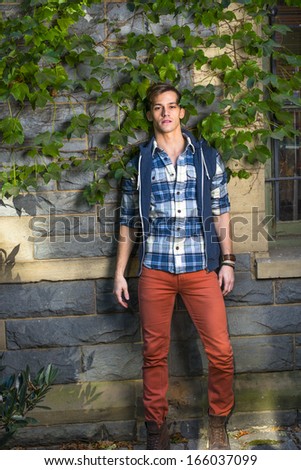 Dressing in a blue and white pattern shirt,  a blue hoody vest,  red jeans, a young guy is standing by a window and against the wall with green ivy leaves. / Portrait of Young Guy