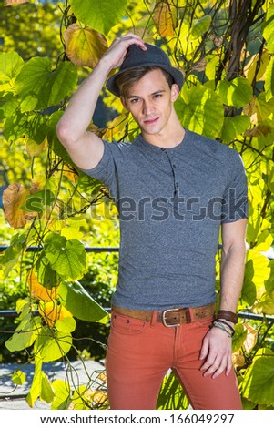 Dressing in a gray long sleeves with roll-tab Henley shirt and red jeans, a young handsome guy is standing by green plants,  wearing a woolen Fedora hat and looking at you. / Portrait of Young Guy