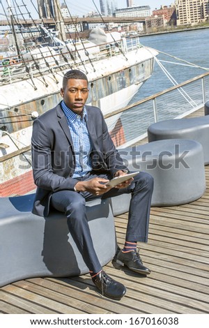 A young black businessman is sitting on a deck,  reading on a tablet computer. The background is a harbor. / Working Outside