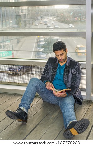 Against a glass wall, a young guy with beard and mustache is sitting on the floor, reading a book. Background is a busy street scene. / Leisure Time