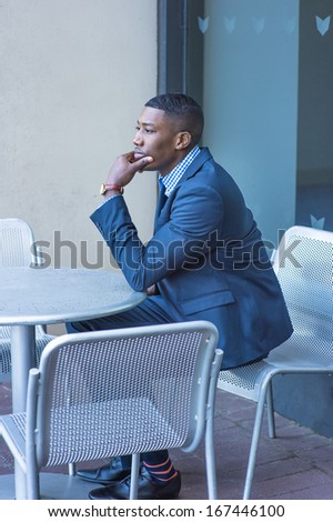 A hand touching his chin, a young black businessman is sitting on a chair by a table outside, thinking. / Thinking Outside