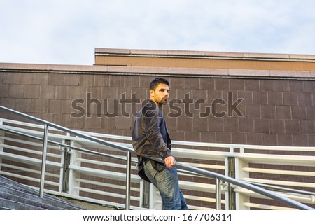 Dressing in dark purple woolen blazer and jeans, a young guy with beard and mustache is standing against a metal railing on stairs, under sunshine of sunset, thinking. / Thinking Outside
