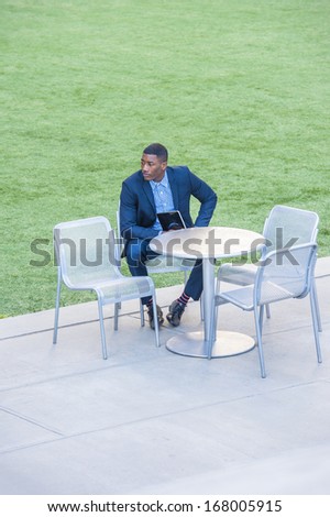 A young handsome black businessman is sitting on a chair by a green lawn, holding a small computer, looking around and waiting for you. /Waiting for you