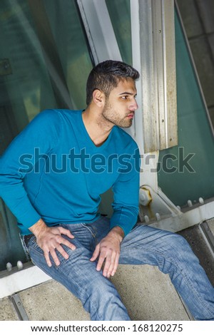 Dressing in a blue sweater and jeans , a young handsome guy with beard and mustache is sitting on the corner by a structure, bending over, looking down, lost in thought. / Deeply Thinking