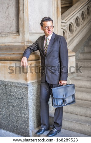 Dressing formally in a suit, a tie and leather shoes, wearing glasses, carrying a leather bag,  a young businessman is standing on stairs outside an office, looking away and waiting for you.