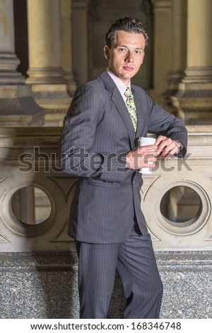 Dressing formally, holding a cup of coffee, a young businessman is standing in an office hallway, seriously looking at you. / Taking Break